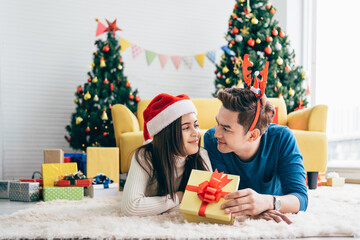Fototapeta na wymiar Young happy Asian woman wearing a Santa Claus hat with her boyfriend with a Christmas gift while lying down on the carpet and looking at the camera with a Christmas tree in the background.