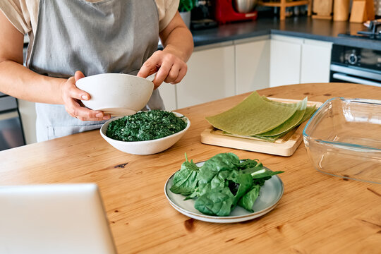 Una donna cucina lasagne fatte in casa di spinaci e ricotta da una ricetta che ha trovato online sul suo laptop. Piatto della tradizione italiana. Messa a fuoco selettiva.