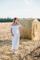 Naklejka na ściany i meble beautiful pregnant brunette in a white dress near the haystacks in the field at sunset. High quality photo