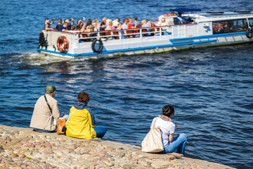 People sitting by the river look at a passing pleasure boat