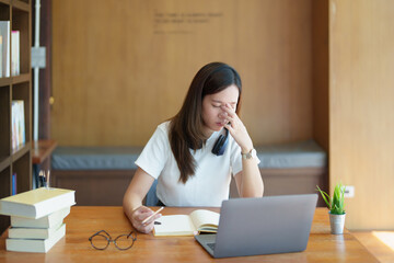 A portrait of a young Asian woman using a computer, wearing headphones and using a notebook to...