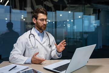 Online consultation of mature serious doctor with beard, man in medical gown using laptop for video call, explaining treatment instructions to patient, working inside modern clinic in the evening.