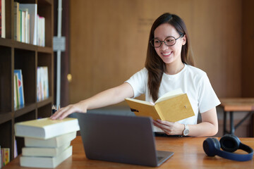 Portrait of a teenage Asian woman using a computer and notebook to study online via video conferencing on a wooden desk in library