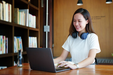 Portrait of a teenage Asian woman using a computer and notebook to study online via video conferencing on a wooden desk in library