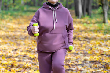 Unrecognizable pensioner woman performs exercises with small dumbbells in the park. Running with dumbbells in place. Outdoor fitness classes.