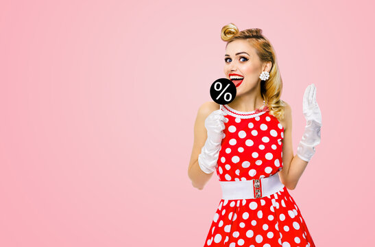 Very tasty discounts, rebates, deals concept image. Cheerful beautiful woman licking signboard with % sign, dressed in pinup red dress, isolated on rose pink background. Black Friday sales ad.