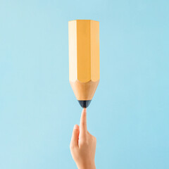 A huge yellow wooden pencil standing on the index finger of a child's hand. Pastel blue background. Minimal abstract education, school concept. Handwriting or drawing exercises.