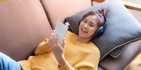 Young beautiful Asian woman laying in sofa and playing smartphone with headphones in home...