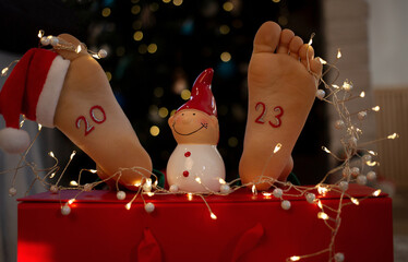 numbers 2023 are written on bare feet of child, small hat of Santa Claus is dressed on toes, around...