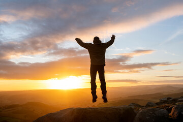 Fototapeta premium silhouette of jumping man with arms raised reaching the destination and on the top of mountain in Peak District at sunset on autumn day Travel Lifestyle concept