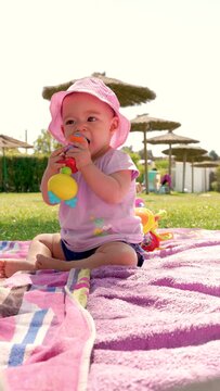 vertical video of a baby sitting with a pink cap on a towel in the pool biting a toy to relieve her gum pain caused by the eruption of the first teeth, 4k video, close-up, slow motion
