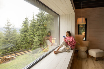 Woman sits with phone on window sill and enjoys scenic view on mountains while resting in wooden...