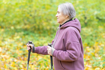 Gray-haired woman with Scandinavian walking sticks. Side view. Autumn background with copy space.