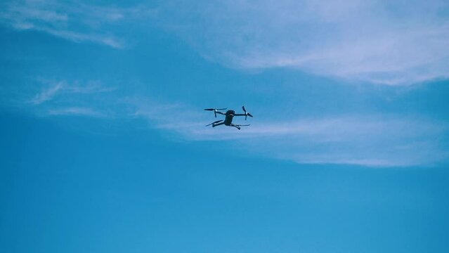 The drone flies against a background of blue sky and clouds, a view from below. The quadcopter with the camera hovers at a low altitude. Fast-rotating drone propellers.