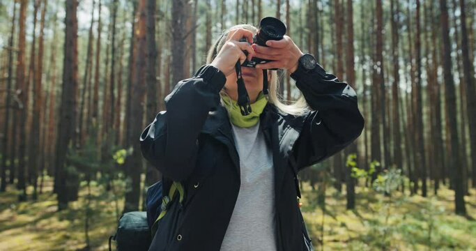 Satisfied young woman with headband wearing sports uniform standing in the middle of forest with travel backpack behind and mat holding modern camera in hands taking pictures of nature while relaxing.