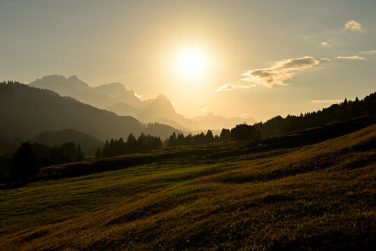 Die Sonne geht über dem Wetterstein Massiv mit der Zugspitze unter und im Vordergrund sind die typischen Buckelwiesen der bayerischen Voralpen zu sehen.