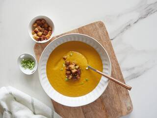 Top view of vegetable puree with pumpkin on plate with spoon on wooden board and marble kitchen countertop. Bread croutons and spring onion garnised.