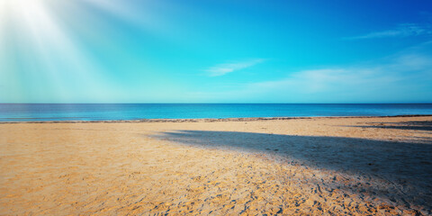 Bright long sunrays illuminate empty sandy beach with calm sea and clear blue sky