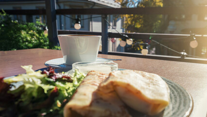 Cup of coffee or tea and plate with pancakes and salad standing on wooden street cafe table outside...