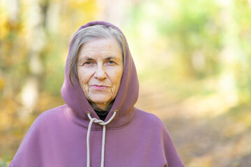 Portrait of a contented gray-haired pensioner in a hood. Purple hoodie. Against the backdrop of a forest in bokeh with space for copy.