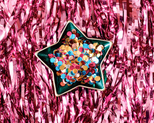 Multicolored glittery confetti are in a festive star-shaped plate. The background is made of tinsel. Minimalistic template for christmas, new year. Flat lay.