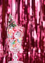 The concept of counting down to the new year. A toy Christmas tree and a small alarm clock, along with multi-colored dragees, are in a vintage glass goblet against a background of tinsel.
