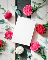 An open blank notepad is above aster flowers and eucalyptus branches on a white wooden background. Autumn, spring, summer concept of business planning and freelancing. Flat lay. Copy space.