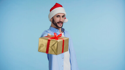 bearded man in red santa hat giving wrapped christmas present isolated on blue
