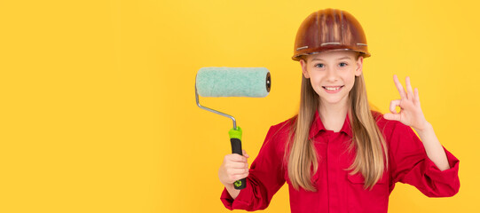 Builder teenager girl in helmet. happy teen child in builder hard hat with paint roller on yellow wall. ok. Child builder in helmet horizontal poster design. Banner header, copy space.