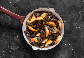 Fried chicken liver with onions and apples in a ceramic frying pan on a dark background, top view