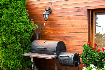 iron black rusty barbecue near wooden house with retro lantern on wall made of boards, cottage with thuja bushes and red pelargonium flowers, place to relax and cook meat and vegetables on grill. - obrazy, fototapety, plakaty