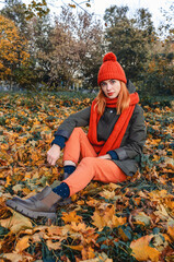 Autumn portrait of a cute girl in an orange warm knitted hat and an orange scarf sitting in a fallen yellow leaf in the park. Romantic look of big eyes. Looks into the camera.