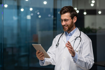 Joyful senior doctor in medical coat using tablet computer for video call and online consultation with patient, man with beard and glasses in medical coat working inside clinic.