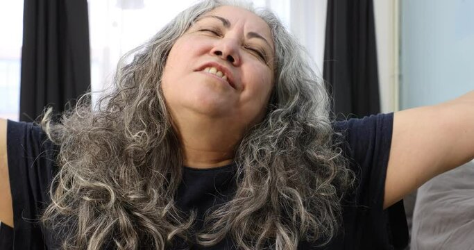 Close-up of newly woken up older woman's face yawning, stretching, making movements with head, cheerful expression to start day with enthusiasm, gray-black long wavy hair. Wellness concept at home