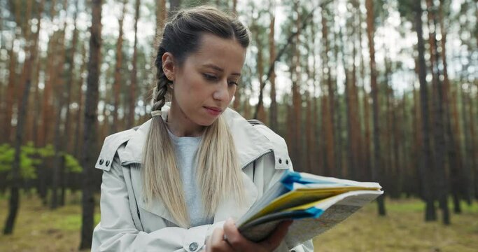 Blonde beautiful woman with braids wearing sportswear standing in the middle of forest holding map searching way home . Family gor lost in forest while trekking hiking doing sports exercises.