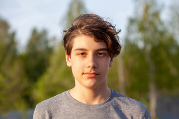 Portrait of a teenager in a grey T-shirt against a background of green foliage. Boy with brown eyes, lit by the setting sun, close-up