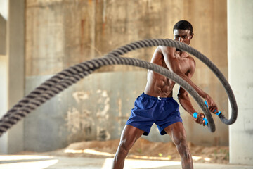 Rope workout. Sport man doing battle ropes exercise outdoor. Black male athlete exercising, doing functional fitness training with heavy rope