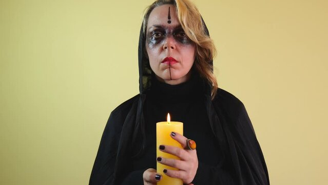 A magic woman in a black cloak with a hood holds a yellow burning candle in her hand on a yellow isolated background. Halloween woman. Fantasy woman portrait