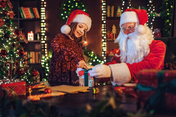 Fototapeta na wymiar Santa Clause packs gifts with his little girl assistant while sitting at the table near fireplace.