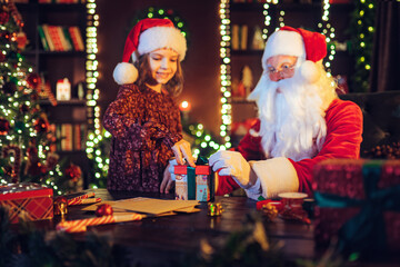 Little girl helps Santa Claus packs giftbox while standing near the table. Selective focus