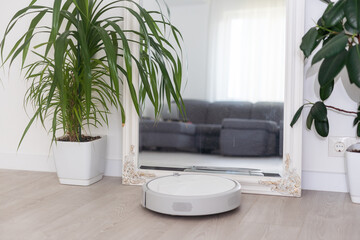 A white robot vacuum cleaner vacuums along the skirting Board near a large window through which the...