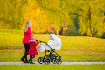Young adult mother and little daughter in red coats pushing white baby stroller and walking at town...