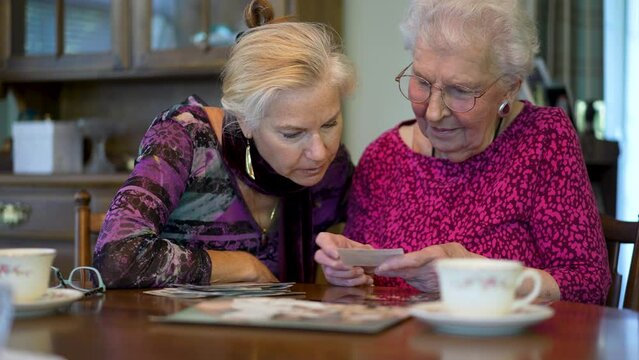 Extreme closeup of senior elderly smiling woman looking at old photos and remembering memories with daughter at the dining room table.