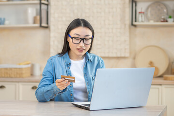 Obraz na płótnie Canvas Online shopping. A young beautiful Asian woman in glasses sits at home at the table with a white laptop, holds a credit card in her hands. Buys and orders goods on the Internet.