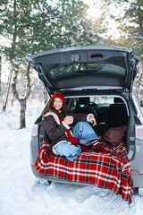 Young woman holds a thermos and drinks tea sitting in car trunk in sunny winter forest. Rest, relaxation, travel, lifestyle concept.