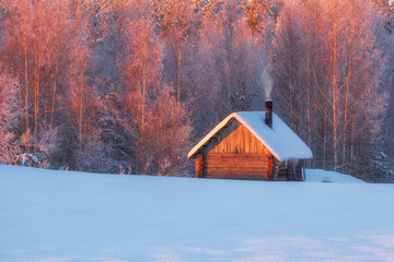 Fairytale small wooden house or sauna near the winter forest in the snow and smoke from the...
