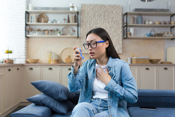 Allergic attack. Asthma. Young Asian woman sitting on sofa at home and using inhaler. Has an asthma...