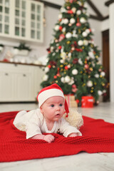 Obraz na płótnie Canvas a baby in a santa hat with a large bubo lies on a red plaid against the background of a Christmas tree.