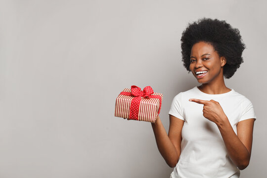 Beautiful happy woman smiling, holding gift box and pointing at red present on white background