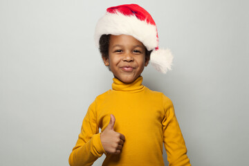 Small optimistic black child boy in Santa hat smiling and showing thumb up on white background